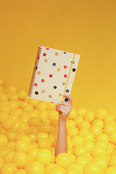 A single hand extending out of a yellow ball pit and holding a cute notebook with colorful dots on a yellow background