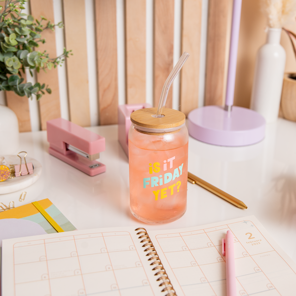 16 oz can glass with lid + straw, text that reads "IS IT FRIDAY YET?" is on the front of the glass in multi-color font. Glass is filled with juice, and surrounded by various stationery supplies on a desk.