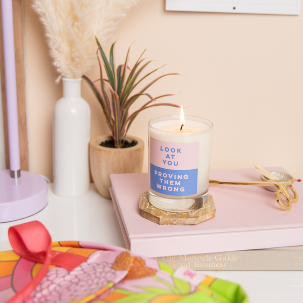 Rocks glass candle with pink and blue label on front with text that reads "LOOK AT YOU PROVING THEM WRONG". Candle is sitting on a wooden coaster and pink book, surrounded by various stationery items.