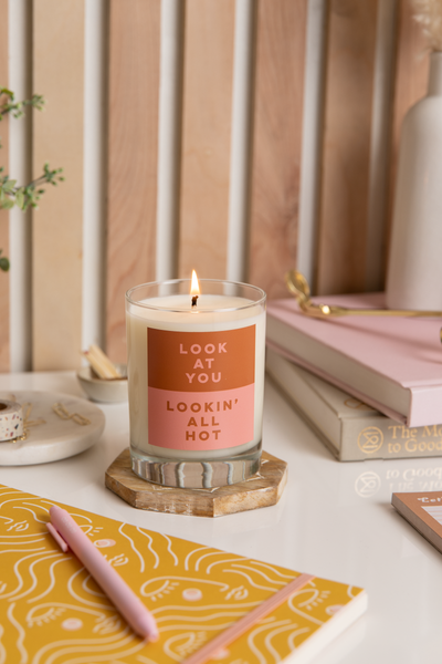Rocks glass candle with a pink and burnt orange colored label on the front that reads "LOOK AT YOU, LOOKIN' ALL HOT". Candle sits on a wooden coaster and is surrounded by various stationery items.