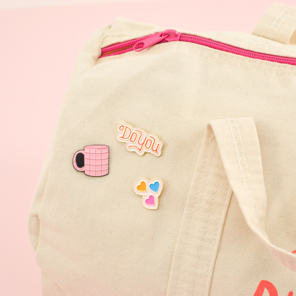 Our Do You, Grid Coffee Mug, and Heart enamel pins pinned to a cute duffel bag.