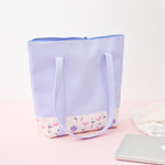 A periwinkle tote bag with magic sprigs on the bottom sitting next to a macbook