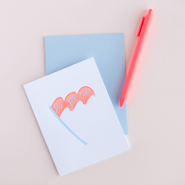 White greeting card with a blue and coral pennant with text "Yes You Can!". There is a blue envelope and a neon coral jotter pen in the background.