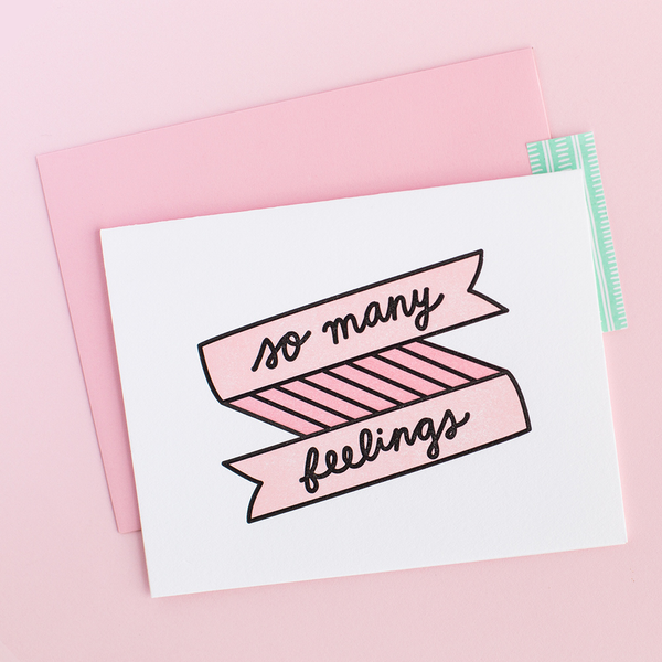 White greeting card with a pink and black banner and scripted writing "so many feelings". There is a pink envelope and a pink background.