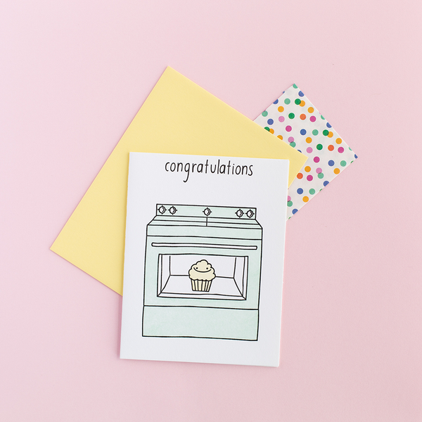 White greeting card with a mint oven. The oven has a clear window with a smiling muffin in it. The text at the top is "congratulations". There is a yellow envelope and a small color polka dot gift bag against a pink background.