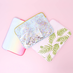 Pile of cute laptop sleeves in Buds, Confetti, and Meltdown sitting on a pink table. Meltdown is pastel rainbow, confetti is clear vinyl and buds is a pink background with green palms all over the bottom half.