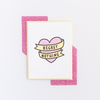 White greeting card with a pink heart and yellow banner, both outlined in black. The banner has the text 