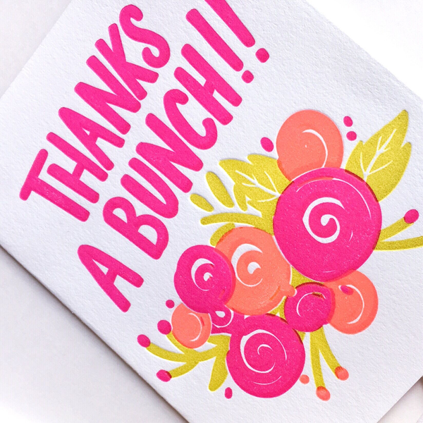 A white greeting card with the text "Thanks A Bunch!!" in bright pink. There are bright pink, orange and yellow flowers and leaves at the bottom. The card is a diagonal and cropped close up photo.