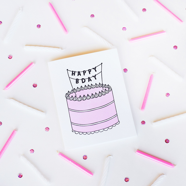 A white greeting card with a pink, black and white birthday cake. On top of the cake is a banner with the text "HAPPY BDAY" in black letters. The background has white and pink candles scattered and pink sequin.