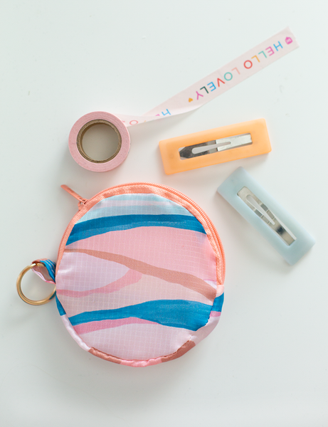 An abstract, wave-like design circle pouch with a zipper and a keyring. The pouch is in pastel and neutral colors. There is also washi-tape and hair clips displayed with the pouch.