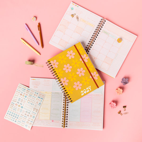 Day to Daisy 2024 Dated Goal Getter Planner laying on an open planner with jotter pens, a "here comes the sun" enamel pin and colorful  flower clips.