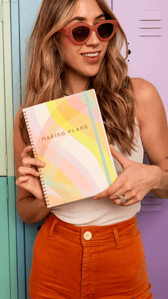 girl with brown sunglasses holding a perpetual planner with pastel multi-color waves with text saying 'making plans' and a bright blue elastic ban to keep the planner closed