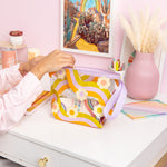 Clear colorful wavy rainbow with daisy's pouch on a desk with misc items. Poster and pink background.
