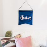 Thrive Wallflower is a cute canvas wall hanging in navy blue with pink lettering above a cute couch with two throw pillows and a fur throw blanket