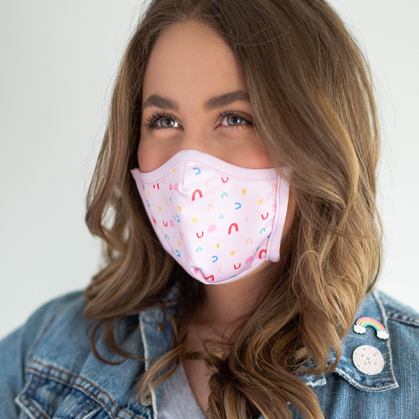 A brunette wears a pink facemask with small rainbow illustrations.