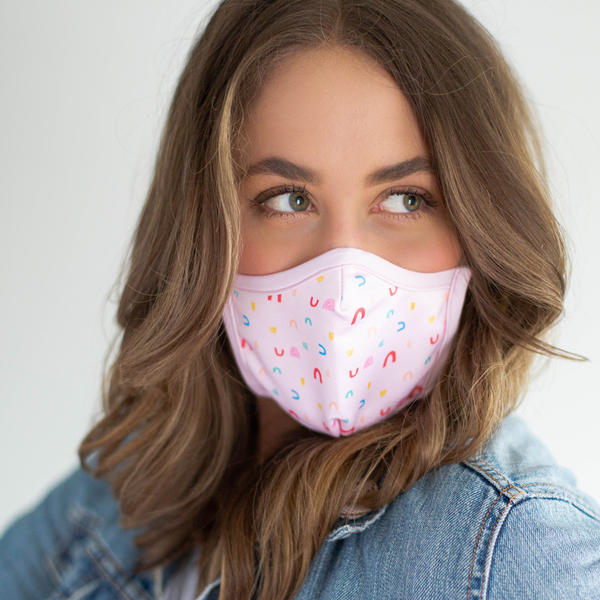 A brunette woman wears a pink facemask with small rainbow illustrations.