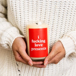 Someone holding a 12 oz. Holiday candle with a red decal that says, "i fucking love presents."