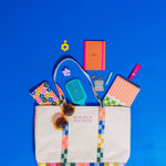 Image contains a large tote with the text, "On My Way To Fuck Shit Up" in a bold checker pattern with dimensions 15" x 13.5" x 5". There are items coming out bag that includes a penny key ring in a floral pattern, sunglasses, a turquoise brush, a key charm that reads "Drink Your Water", a flower claw clip, a small highlighter marker, lip balm, single yellow flower shaped earring, a mini orange notebook, and a taskpad, against a blue background.    