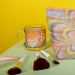 Colorful "dont talk to me" text on a glass mug with smiley faces  on a green surface with misc items and a kaleidoscope pouch and a mustard yellow background