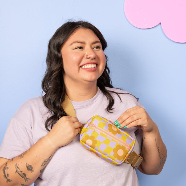 Blue background with a girl smiling wearing a small pastel purple and green checkers with flowers pattern hip bag 