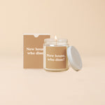 A white, flaming candle that is inside a clear glass jar with the lid leaning up against the lit candle. The glass candle has a light brown sticker on it with the words New house, who diss?" written in a light tan color. There is a light brown box beside the with the same wording on it.