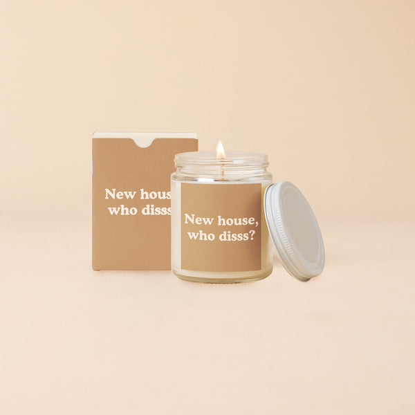A white, flaming candle that is inside a clear glass jar with the lid leaning up against the lit candle. The glass candle has a light brown sticker on it with the words New house, who diss?" written in a light tan color. There is a light brown box beside the with the same wording on it.