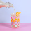 A pink drink being poured into the carnival checker glass tumbler that has lemons on top and ice in it.