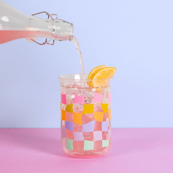 A pink drink being poured into the carnival checker glass tumbler that has lemons on top and ice in it.