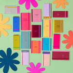 Image contains multiple notebooks, taskpads, and notepads against a light green backdrop and different colored flower props. Large notebooks dimensions are 4.5" W x 8.125" H and mini notebooks dimensions are 4.5" W x 6" H, both with tape binding, and elastic closure. Taskpads dimensions are 3" W x 7" H with coil binding and elastic band closure. Tearaway notepads with gluetop binding and dimensions  3.5" W x 7" H. 