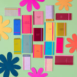 Image contains multiple notebooks, taskpads, and notepads against a light green backdrop and different colored flower props. Large notebooks dimensions are 4.5" W x 8.125" H and mini notebooks dimensions are 4.5" W x 6" H, both with tape binding, and elastic closure. Taskpads dimensions are 3" W x 7" H with coil binding and elastic band closure. Tearaway notepads with gluetop binding and dimensions  3.5" W x 7" H. 