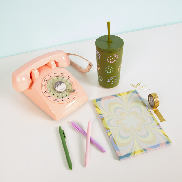 Kaleidoscope pastel color floral notepad with smiley "fuck off" cold cup with misc items on a light blue surface.