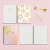 Open daily planner with gold floral pattern and lined weekly spread, next to accessories including pens, washi tape, and a flower clip.
