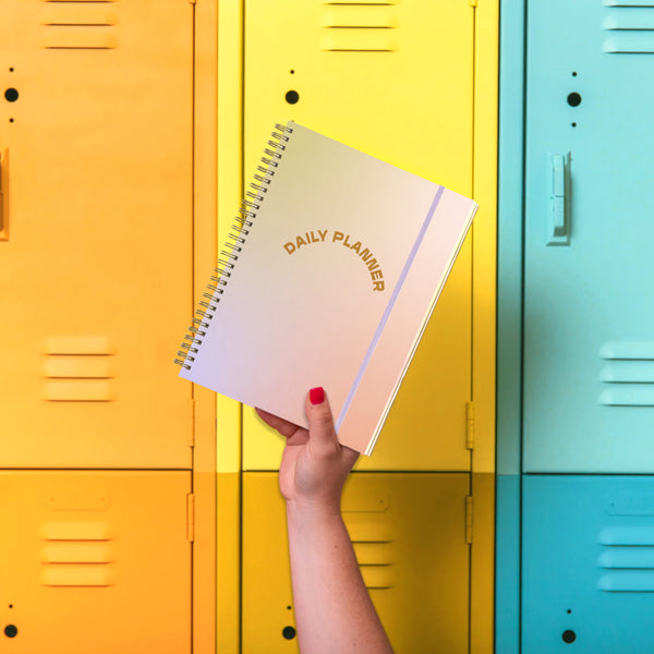 Hand holding a pastel daily planner in front of yellow and turquoise lockers.