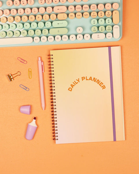 Daily planner with “Daily Planner” text on the cover, placed beside office supplies and a mint-and-peach keyboard on orange background.