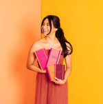 A model standing in front of a yellow backdrop holding multiple notebooks in the colors bright pink, light pink, maroon, and orange.  Larger notebooks dimensions are 4.5" W x 8.125" H , with tape binding, and elastic closure. Mini notebooks are in the dimensions 4.5" W x 6" H, with tape binding, and an elastic closure. 