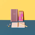 A collage featuring three different notebooks. The first notebook is maroon, has a pink elastic closure  and has "Manifest That Shit"written on the cover. The second notebook has Coil Binding with a pink Elastic Closure and a fun bold colorful rectangular shaped cover. The third coil binding notebook is opened and has a yellow Jotter resting on it. There are three binder clips next to the notebooks. 
