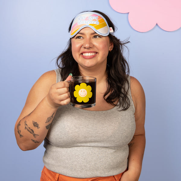 Girl holding a green smiley flower glass mug with sleep mask on her head on a light blue background.