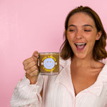 Girl holding a "fuck around and find out" text glass mug with a blue smiley face and small daisys on the each side with a pink background