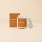 A Yippie Toot Candle Jar with Lid that says Burning it down in pink on a cinnamon orange like background with a matching box. Smells like a fresh woody air. 