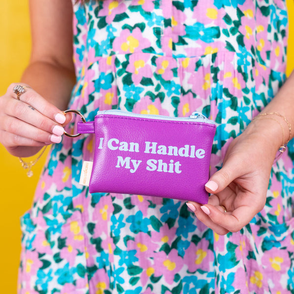 Person holding a purple card pouch with a light blue zippier with light blue test on the pouch that says "I Can Handle My Shit" against a yellow background