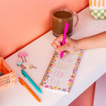 Person writing in a colorful notebook with a pink pen on a desk against a pink wall.