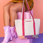 Person holding a tote bag with 'Unbothered' text on the front against a pink background. The Tote is canvas with the straps being maroon and the bottom of the bag is a striped pattern 