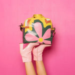 Nostalgic Skate Bag being help up by a women's legs wearing a pair of pink heeled booties. The Skate Bag is a retro flower pattern with greens, pinks, citron, and mint. The background is pink