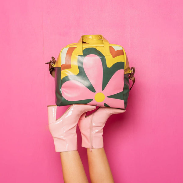 Nostalgic Skate Bag being help up by a women's legs wearing a pair of pink heeled booties. The Skate Bag is a retro flower pattern with greens, pinks, citron, and mint. The background is pink