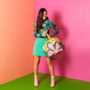 Nostalgic Skate Bag being swung by a women in a teal outfit. The Skate Bag is a retro flower pattern with greens, pinks, citron, and mint. The background is pink, orange, and green.