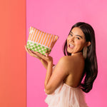 A Women in a ruffle dress and dangle earrings holding up a Tweedle Dee Pouch. The Pouch had pink and orange stripes and at the bottom there is a green and floral checkered print pattern. Between the stripes and checkers there is a light pink scallop to separate them. 
