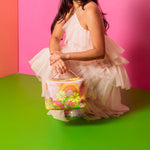 Woman in a white dress holding a retro floral colorful bag against a pink and green background. The bag is the perfect size top use as a cosmetic bag. The pattern is oranges, yellows, greens, and pinks, that make up and floral and rainbow retro design. 