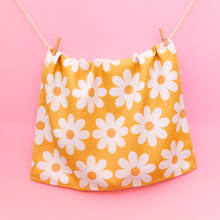 Yellow kitchen towel with a white daisy pattern hanging on a string with a pink background