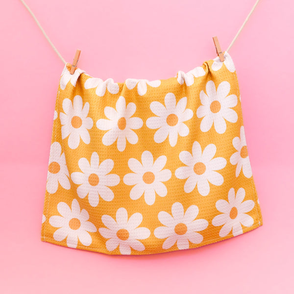 Yellow kitchen towel with a white daisy pattern hanging on a string with a pink background