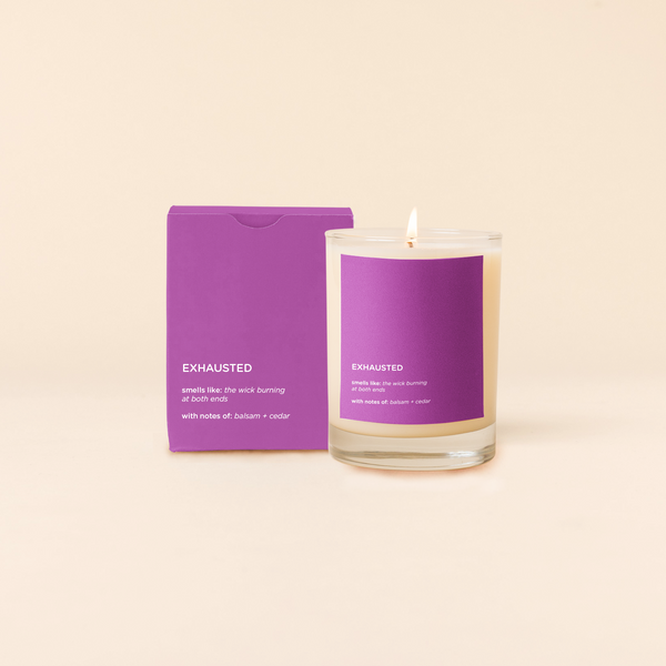 A lit 14oz rocks glass candle with the word "Exhausted" written on a purple sticker. There is a matching box in the purple color with "Exhausted" written on it. The candle is all natural soy-based wax (paraffin-free)!! 